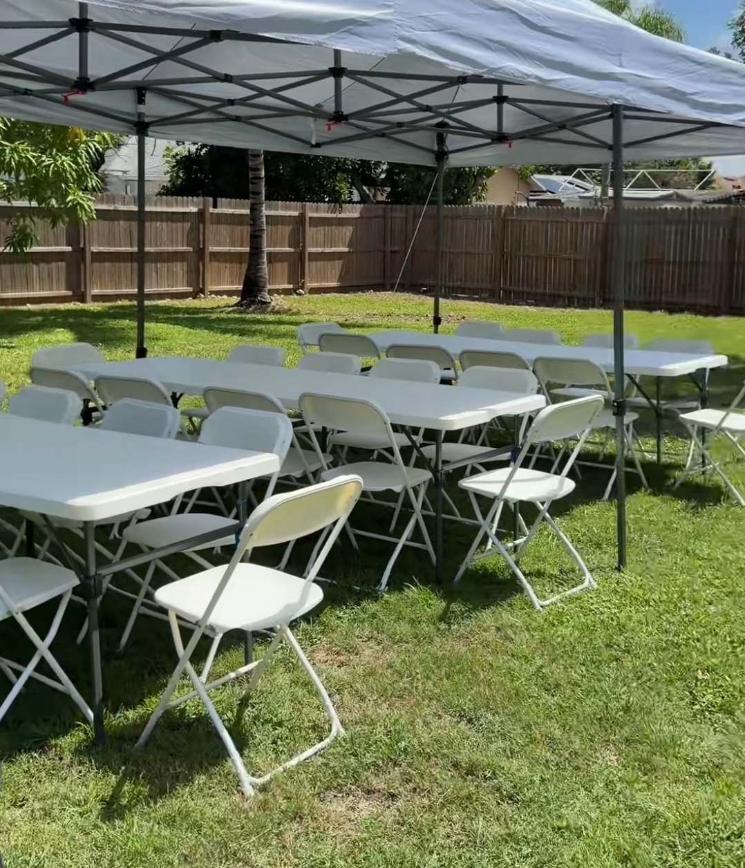 White pop-up canopy over rectangular tables and folding chairs in a fenced backyard